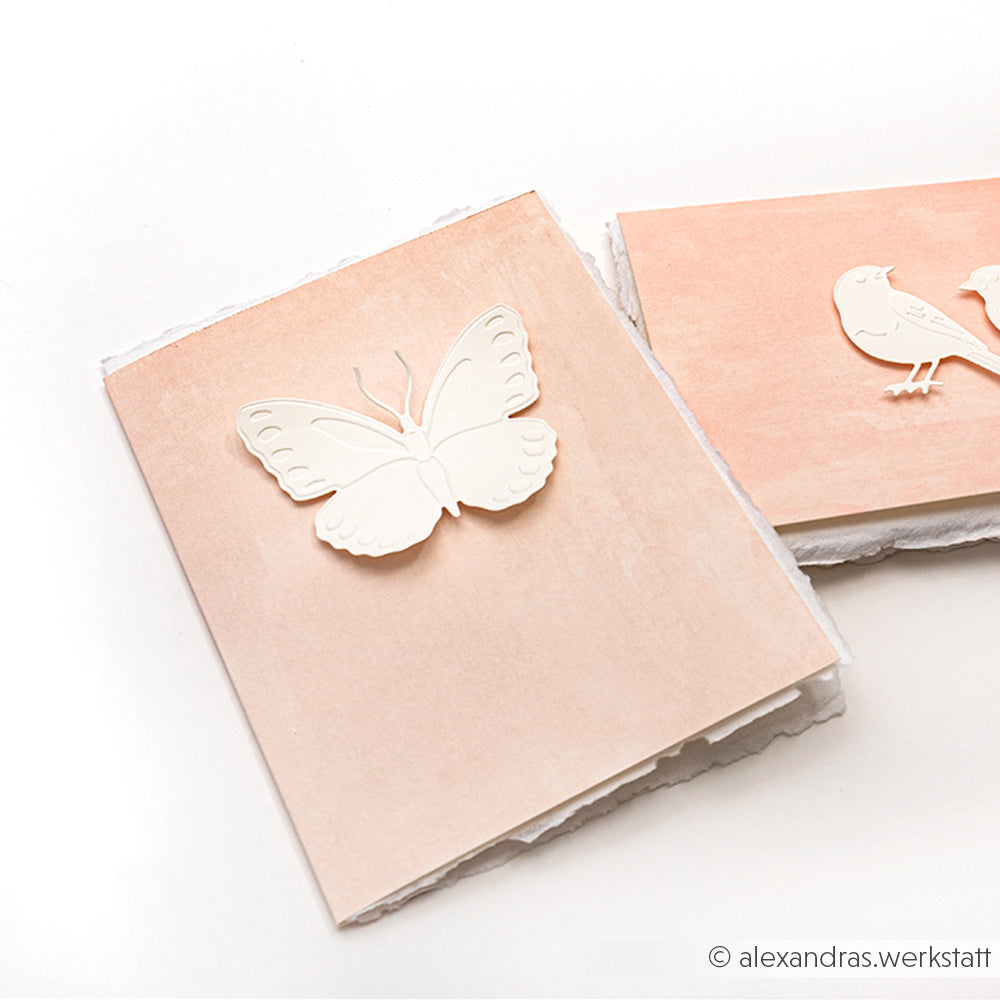 Zwei pastellfarbene Grußkarten von Alexandra Renke, hergestellt mit Stanzsystemen und Cut & Emboss Folder, zeigen zarte Designs: eine mit einem Schmetterling und die andere mit zwei Vögeln, beide auf einem weißen Hintergrund.