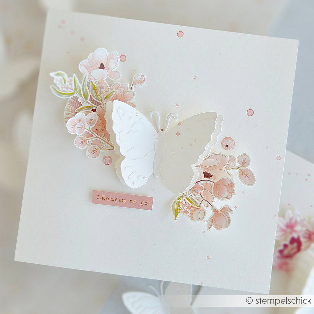 Quadratische Karte mit einem weißen 3D-Schmetterling, der mit der Cut & Emboss-Prägevorlage von Alexandra Renke gestaltet wurde, verziert mit rosa Blumen und dem deutschen Spruch „Lächeln to go“ auf einem zarten Hintergrund – ideal, um mit Ihren Lieblings-Stanzsystemen von Alexandra Renke Schmetterlingsfreuden zu verbreiten.