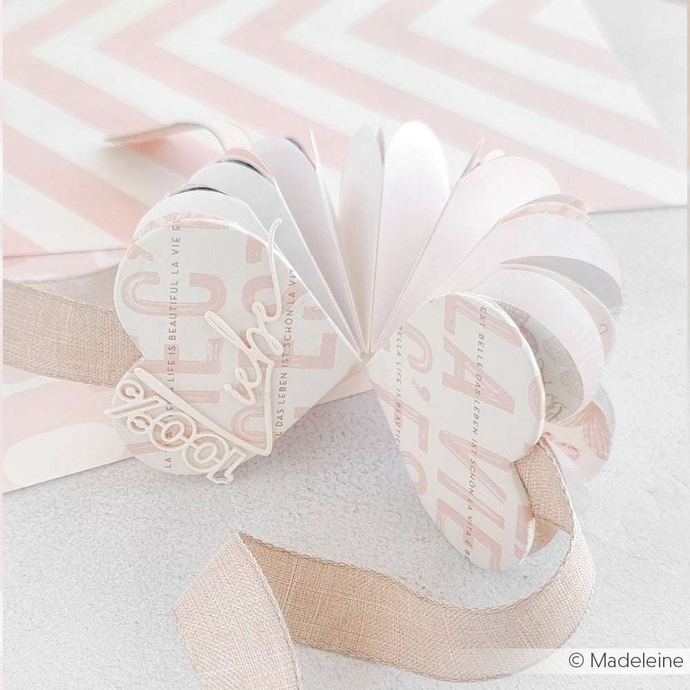 Handgefertigte Papierkarte in Schmetterlingsform von Alexandra Renke mit beigem Band und Stahlbandstanzen auf einem rosa und weiß gemusterten Hintergrund.