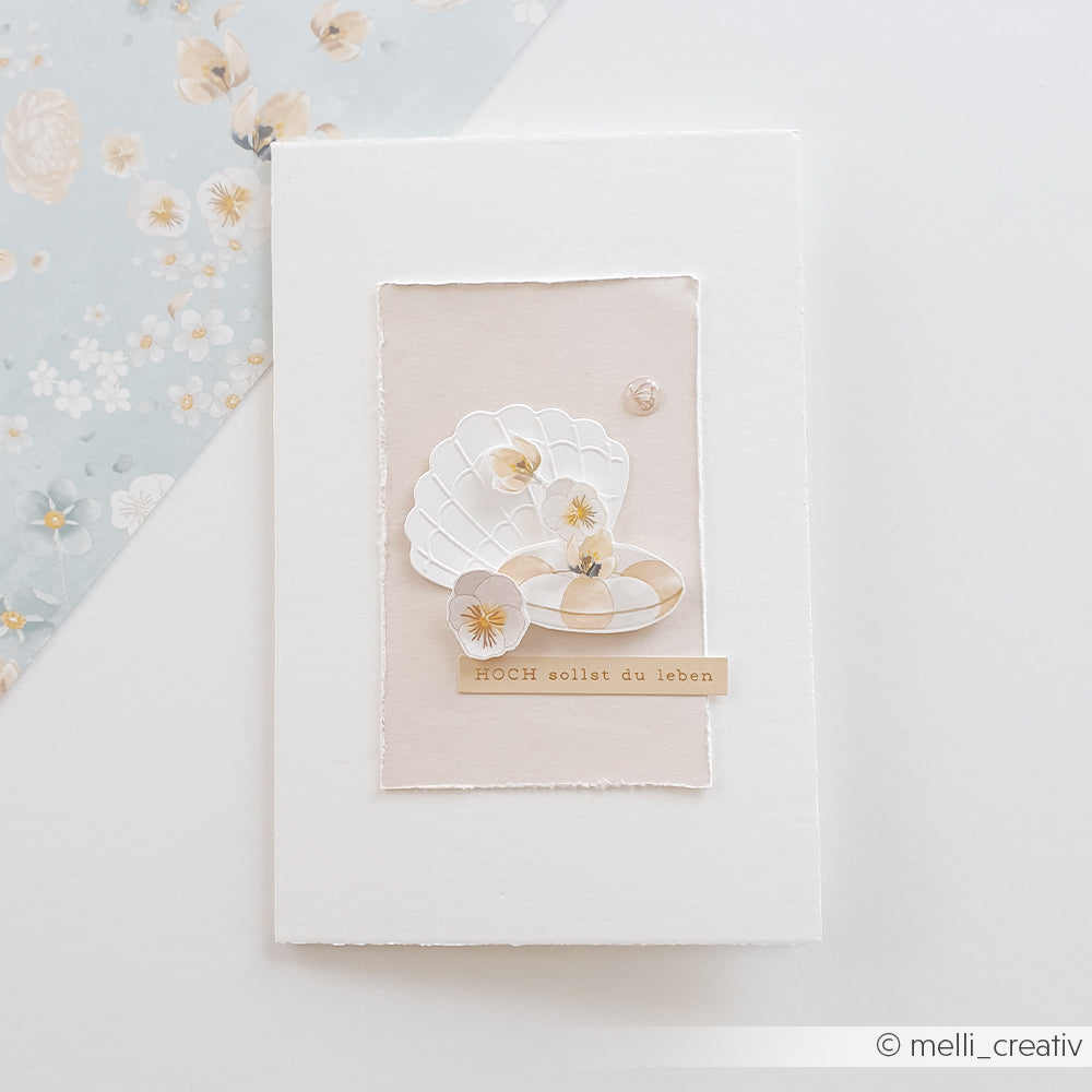 Handgefertigte Karte von Alexandra Renke mit weißen und beigen Muscheln, zarten Blumen und deutschem Text auf strukturiertem Hintergrund, gestaltet mit einer Stanz- und Prägevorlage für filigrane Details.