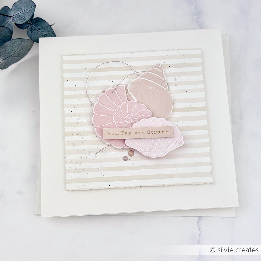 Quadratische Karte mit pastellfarbenen Muscheln und dem Schriftzug „Ein Tag am Strand“ auf gestreiftem Hintergrund, entworfen von silvie.creates für Alexandra Renke. Diese Karte eignet sich perfekt für Sommerprojekte und bringt die zarten Muscheln, die mit Stanzsystemen und einer Cut & Emboss-Prägevorlage gefertigt wurden, wunderschön zur Geltung.