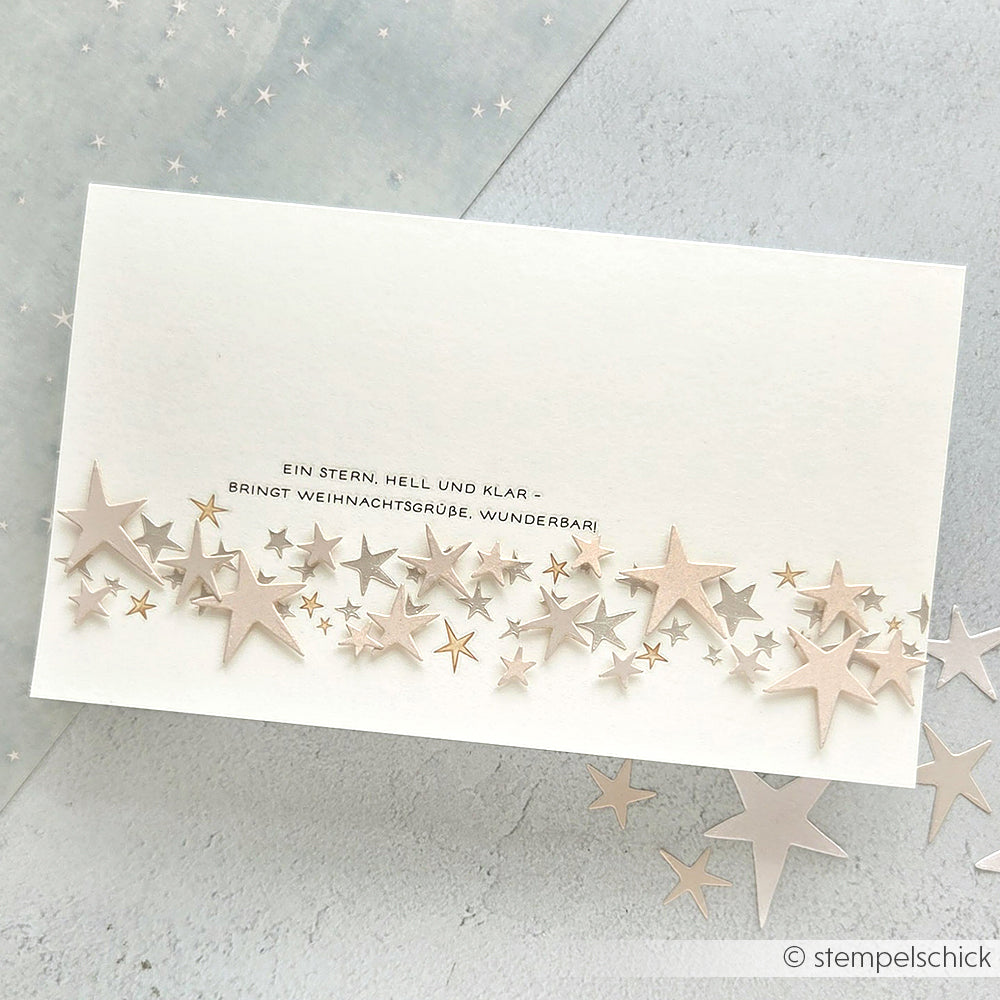Weiße Weihnachtskarte mit verstreuten silbernen und goldenen Sternen, die mit den wiederverwendbaren Schablonen von Alexandra Renke Sternenhäuser Schablone gestaltet wurden, sowie einem deutschen Weihnachtsgruß in der Mitte.