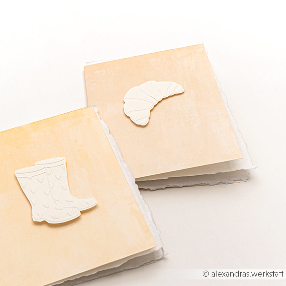 Zwei quadratische Karten von Alexandra Renke mit geprägten weißen Ausschnitten - Regenstiefel auf der einen, Hörnchen auf der anderen - hergestellt mit einem Cut & Emboss Folder. Perfekt, um Ihrer nächsten Kaffeepause auf einem hellen Hintergrund Charme zu verleihen.