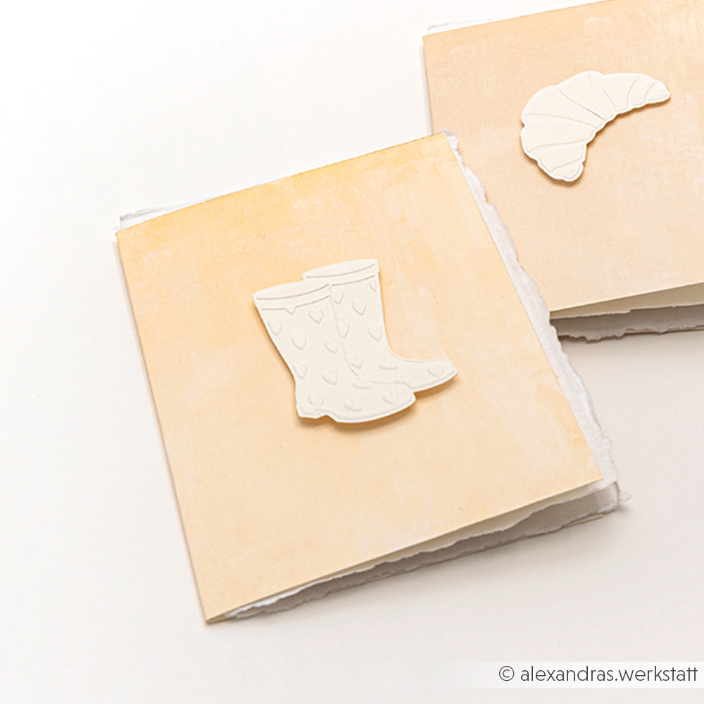 Zwei beigefarbene Karten von Alexandra Renke, die mit dem Cut & Emboss Folder Stanzsysteme gestaltet wurden, zeigen geprägte Regenstiefel und ein Hörnchen auf weißem Untergrund - ideal für kreative Projekte, die von der Gartenzeit inspiriert sind.