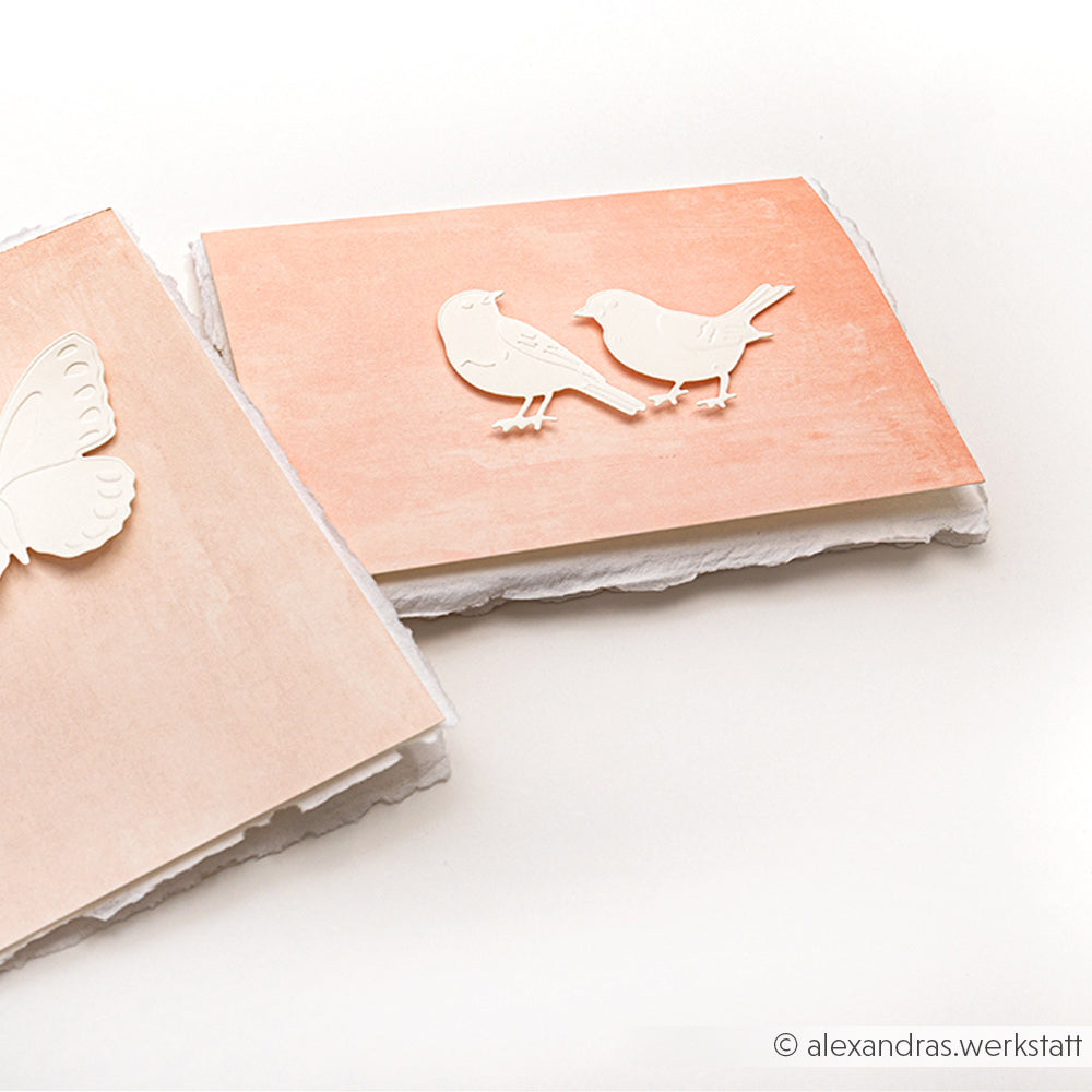 Zwei handgefertigte Karten mit dem Cut & Emboss Folder Stanzsysteme von Alexandra Renke zeigen geprägte weiße Vogel- und Schmetterlingsmotive auf pastellfarbenem Hintergrund - perfekt für die Gartenzeit.