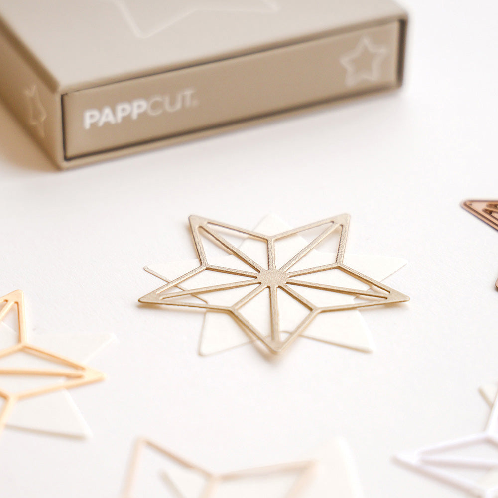 Goldfarbene sternförmige Büroklammer von Alexandra Renke, bekannt als PAPPcut Stern, ausgestellt auf einer weißen Fläche mit anderen Klammern und einer beigen PAPPCUT-Box im Hintergrund - ideal für Buchbinderei oder kreative Projekte.