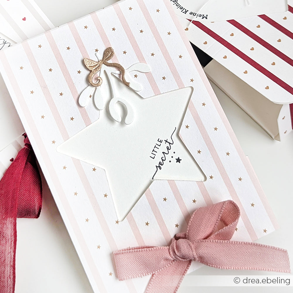Die Grußkarte von Alexandra Renke hat einen PAPPcut Sternausschnitt, rosa Streifen, goldene Punkte, Buchbinden-Details und eine rosa Schleife am unteren Rand.