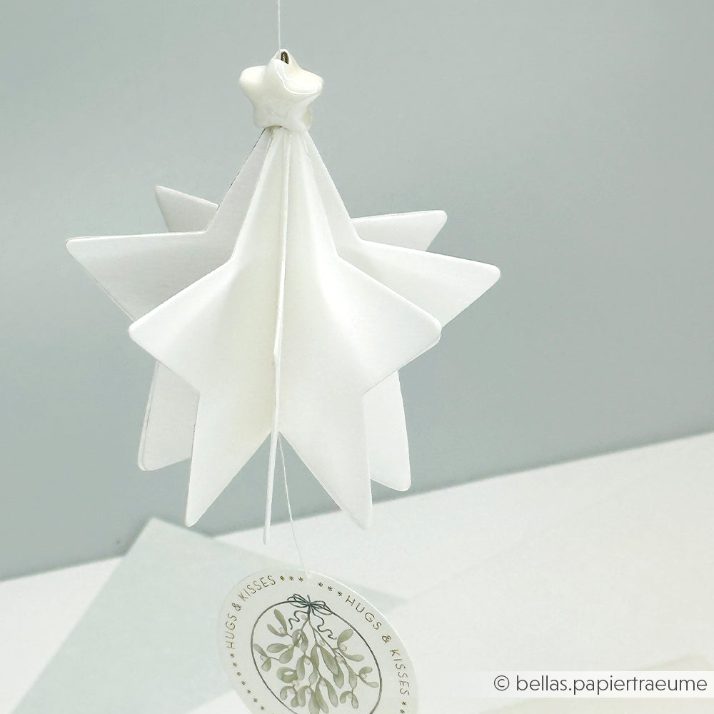 Ein weißes PAPPcut Stern Ornament aus Papier von Alexandra Renke hängt über einer weißen Fläche, die mit einem Hugs & Kisses Anhänger verziert ist.