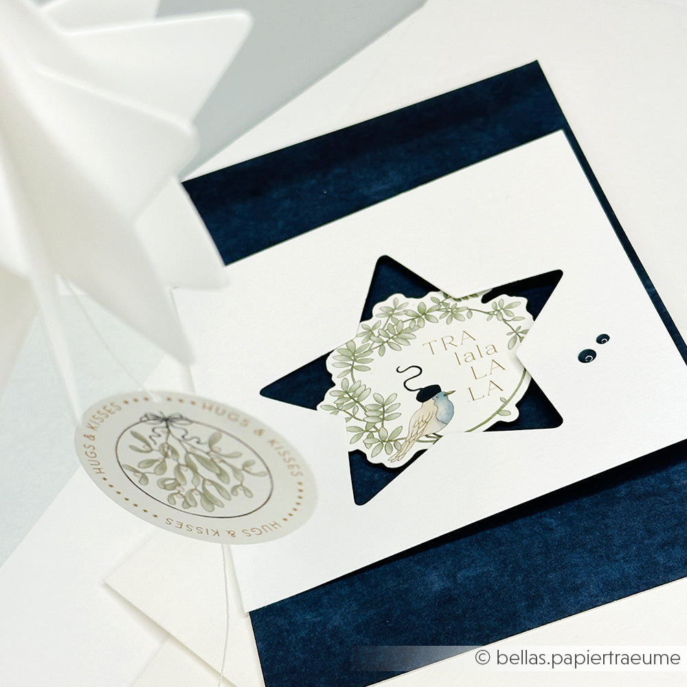 Handgefertigte Karte von Alexandra Renke mit einem PAPPcut-Sternausschnitt, einem Blumenanhänger und dem Text "Tra la la la la" in eleganten Weiß- und Marineblautönen.