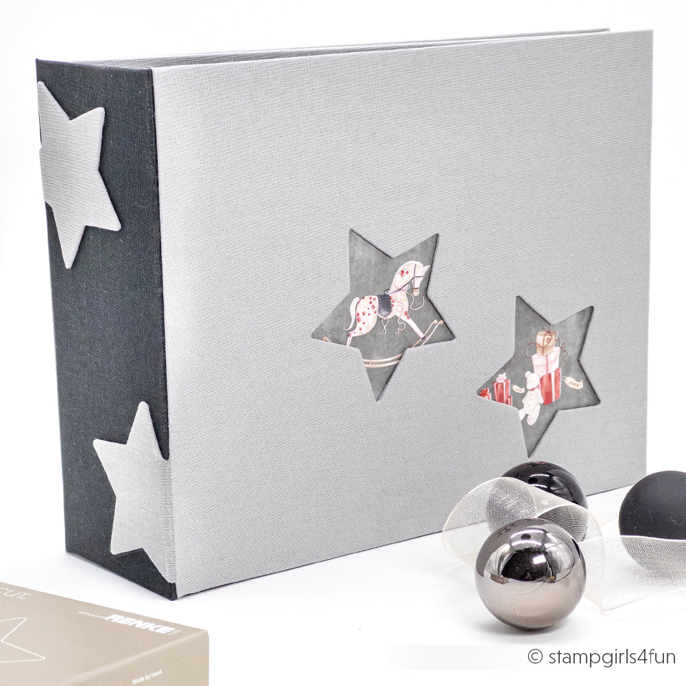 Das grau-schwarze Fotoalbum von Alexandra Renke mit sternförmigen Ausschnitten, gefertigt aus Buchbinden und Stahlbandstanzen, liegt neben silbernen Ornamenten auf einer weißen Fläche.