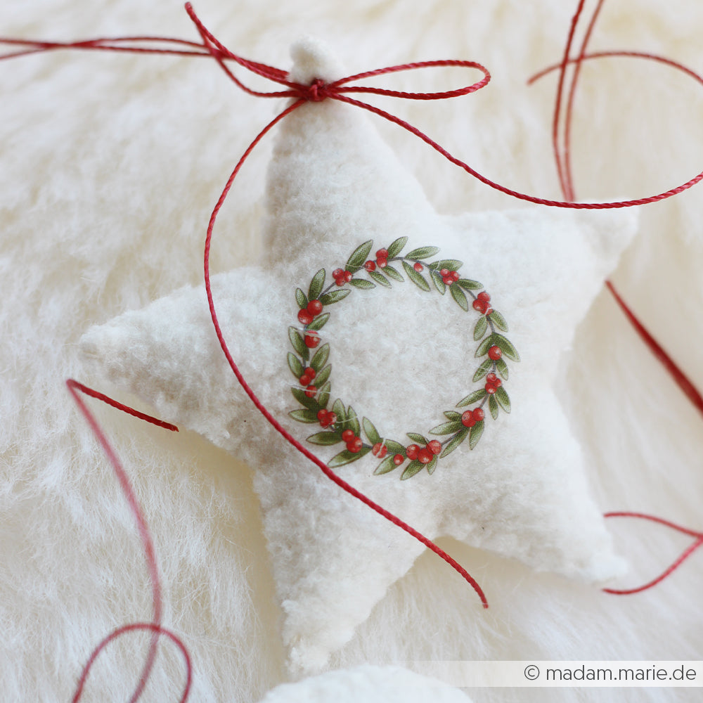 Ein weißes Stern-Ornament von Alexandra Renke mit einem gestickten Kranz und einer roten Schnur, die in Buchbindentechnik gefertigt wurde, ruht auf einer weichen weißen Oberfläche.