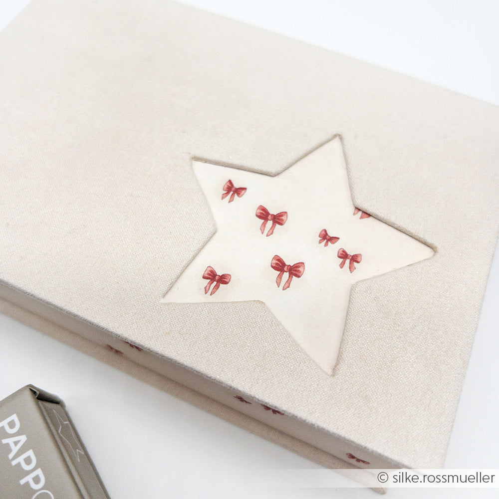 Eine beigefarbene Schachtel von Alexandra Renke ist mit einem sternförmigen Ausschnitt versehen, der im Inneren ein Muster aus kleinen roten Schleifen offenbart, die mit präzisen Buchbinden und innovativen PAPPcuts-Techniken hergestellt wurden.