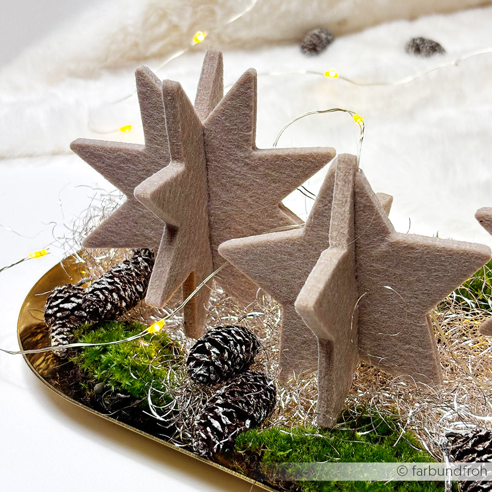 Alexandra Renke hat Sterndekorationen aus Filz, wie PAPPcut Stern und Stahlbandstanzen, auf einem Tablett mit Tannenzapfen, Moos, Lametta und Lichterketten arrangiert.