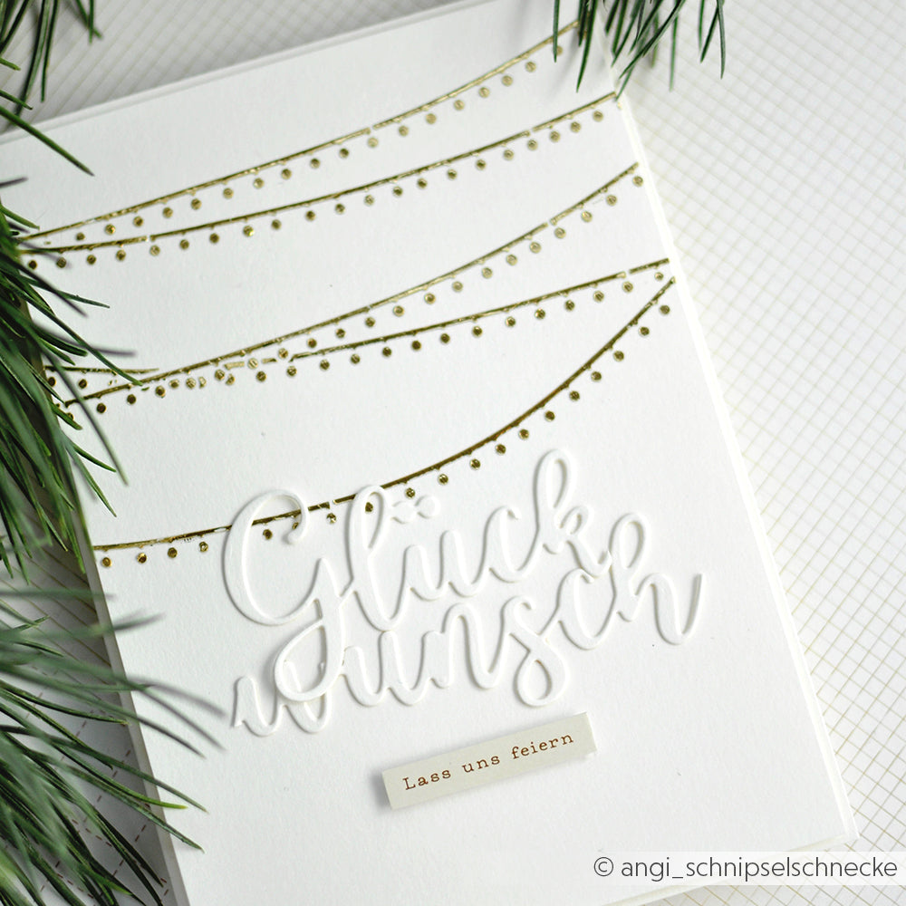 Weiße Karte von Alexandra Renke mit goldener Schablone Lichtergirlanden, erhabenem "Glückwunsch"-Text und einem "Lass uns feiern"-Label, akzentuiert durch Tannenzweige - ideal für Schablonen Papierprojekte oder jeden festlichen Anlass.
