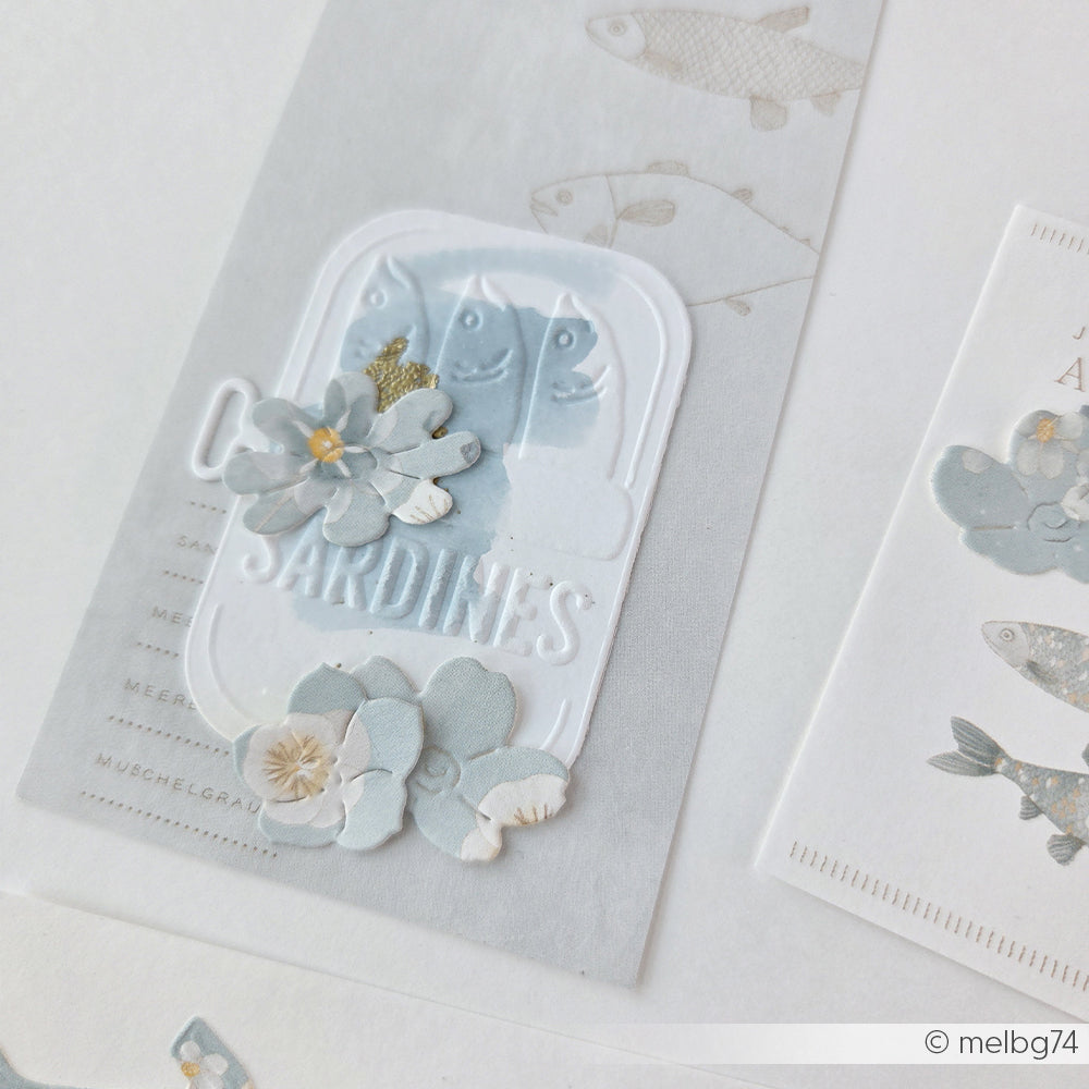 Handgefertigte Karte von Alexandra Renke mit Illustrationen von blauen Sardinen, Blumen und Fischen auf hellem Hintergrund, gestaltet mit „Cut & Emboss Folder“-Techniken für mehr Struktur und Detailreichtum.