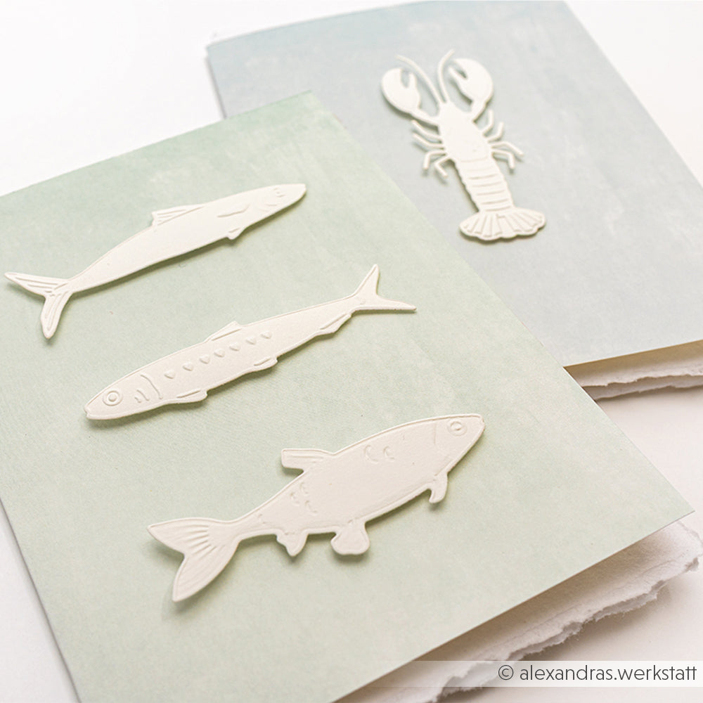 Drei geprägte Fische und ein Hummer, die mit dem Alexandra Renke Cut & Emboss Folder hergestellt wurden, verschönern pastellfarbene Karten auf hellem Hintergrund.