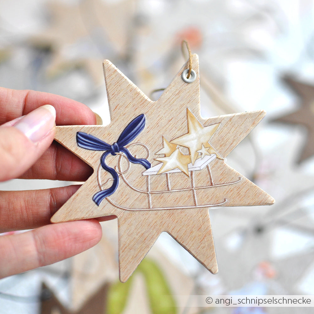 Eine Hand hält ein Alexandra Renke Stern-Ornament aus Holz mit einer blauen Schleife, goldenen Sternen und einem Schlittenmotiv, das mit Präzision im PAPPcut-Verfahren hergestellt wurde.