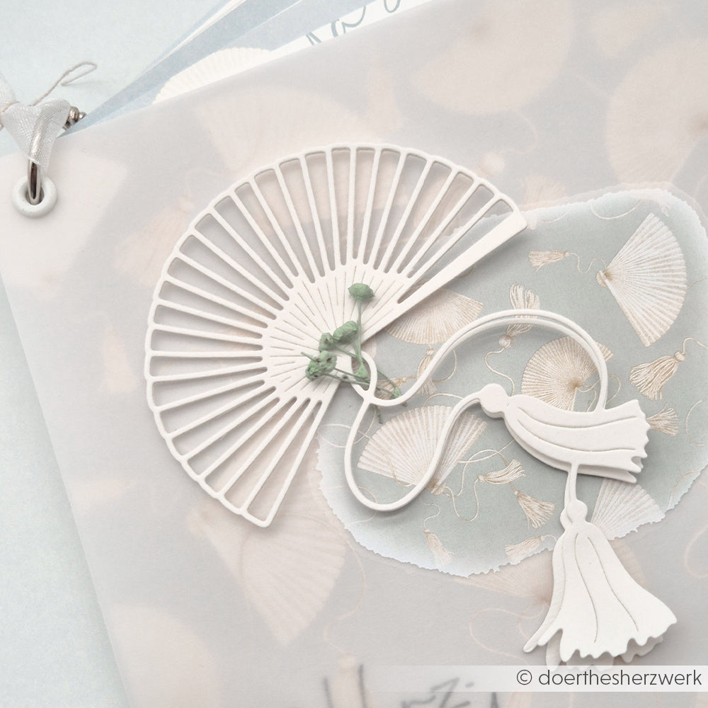 Ein zarter weißer Papierfächer mit Quasten von Alexandra Renke auf einem pastellfarbenen Muschelmuster-Hintergrund - ideal als charmantes Motiv für Bastelprojekte oder zum Verzieren mit Rub-On Transferfolien.