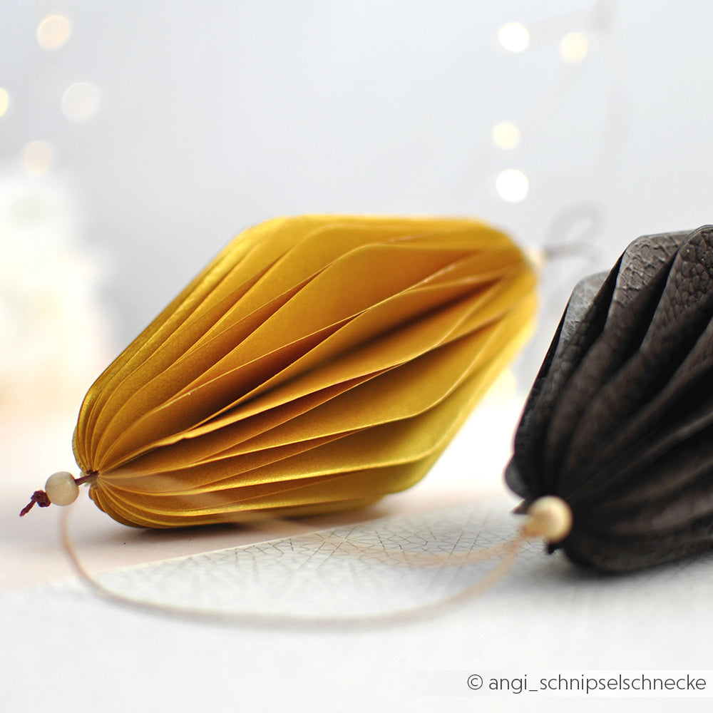 Zwei wabenförmige Papierornamente von Alexandra Renke, die mit PAPPcut-Präzision hergestellt wurden - ein gelbes und ein schwarzes - liegen auf einer weißen Fläche mit verschwommenen Lichtern im Hintergrund.