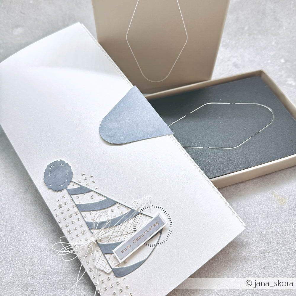 Weiße Geschenkkarte von Alexandra Renke mit einem blauen Partyhut, der teilweise mit einer Schnur aus einer beigen Schachtel gezogen wird - mit PAPPcuts-Techniken hergestellt, ideal für Buchbinden-Fans.