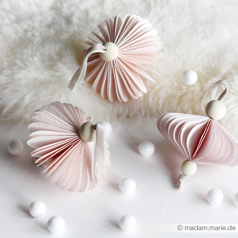 Drei pastellrosa Papierornamente von Alexandra Renke, die mit PAPPcuts hergestellt und mit weißen Perlen und Bändern verziert wurden, ruhen auf einer flauschigen weißen Oberfläche, die mit kleinen runden Verzierungen versehen ist.