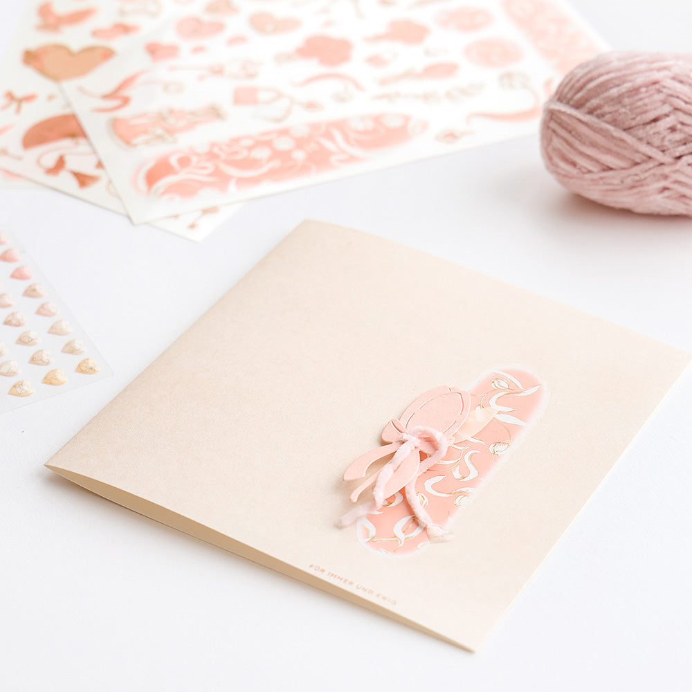 Quadratische Grußkarte von Alexandra Renke mit rosa Schleife und Herzmotiv, umgeben von Aufklebern, einer rosa Garnkugel und kreativen Motiven, die mit Rub-On Transferfolien hergestellt wurden.