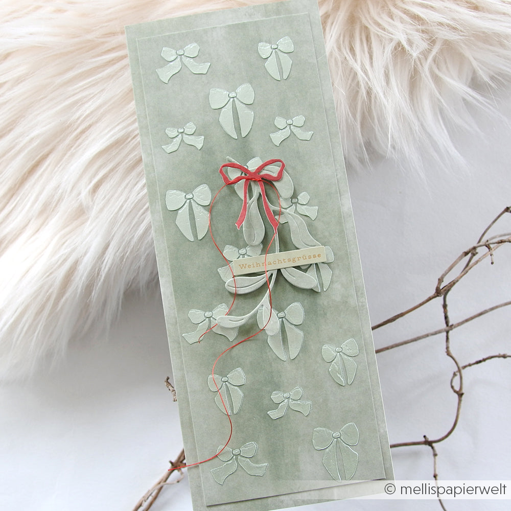 Grüne Grußkarte mit weißen Schleifen und einer rot umrandeten Schleife, hergestellt mit der Schablone Schleifenregen von Alexandra Renke; dargestellt auf einem hellen, strukturierten Hintergrund - ideal für kreative Papierprojekte.