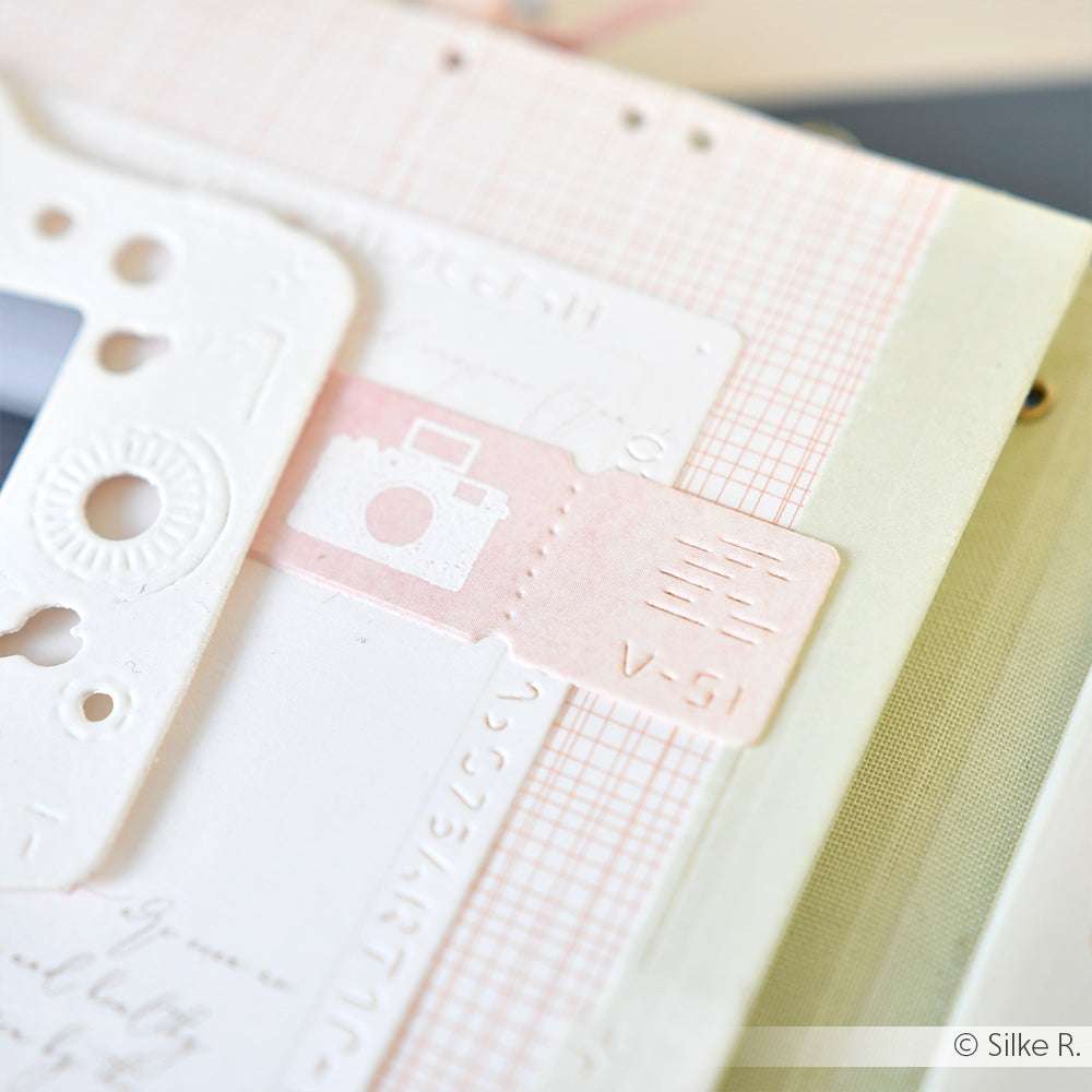 Nahaufnahme von geschichtetem Scrapbook-Papier mit einer rosafarbenen Eintrittskarte mit einem Kamera-Symbol und geprägten Details, perfekt für die Kombination mit Alexandra Renke Photopolymer-Stempeln oder Klarsichtstempeln auf Ihrem Acrylblock.