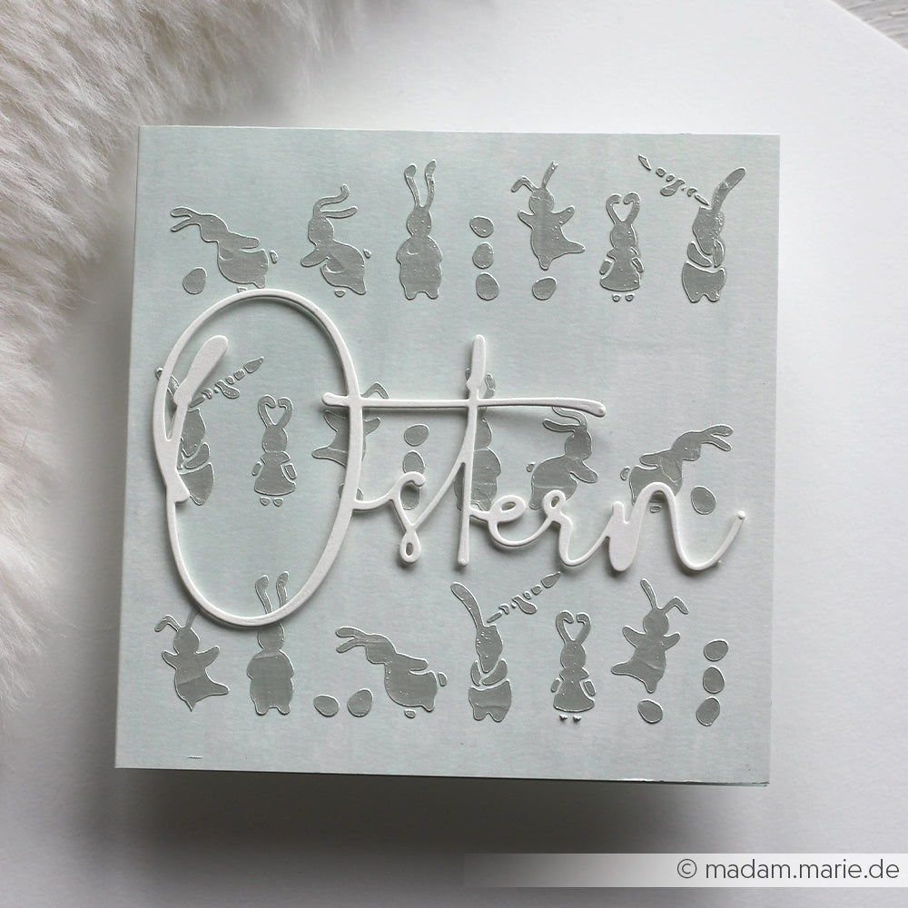 Quadratische Karte von Alexandra Renke mit verspielten grauen Osterhasen und Eiern, "Ostern" in eleganter weißer Schrift und charmanten Schablonenmustern für einen festlichen Touch.