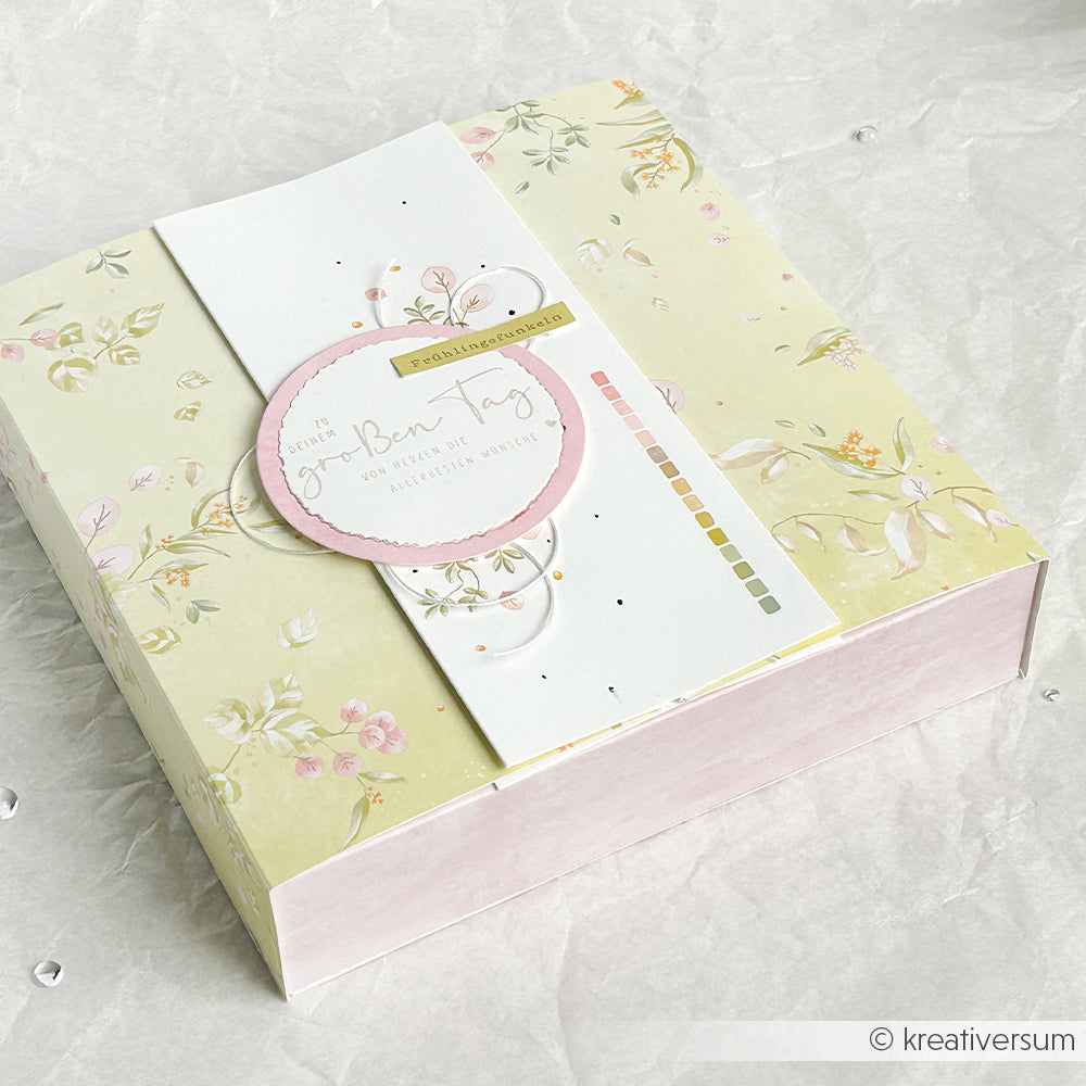 Die pastellfarbene Blumen-Geschenkbox von Alexandra Renke hat ein rundes, rosafarbenes Etikett mit deutschem Text, ideal für Bastelprojekte-Motive oder zum Aufbringen von Rub-On Gartenzauber-Motiven, abgebildet auf leichtem Knitterpapier.