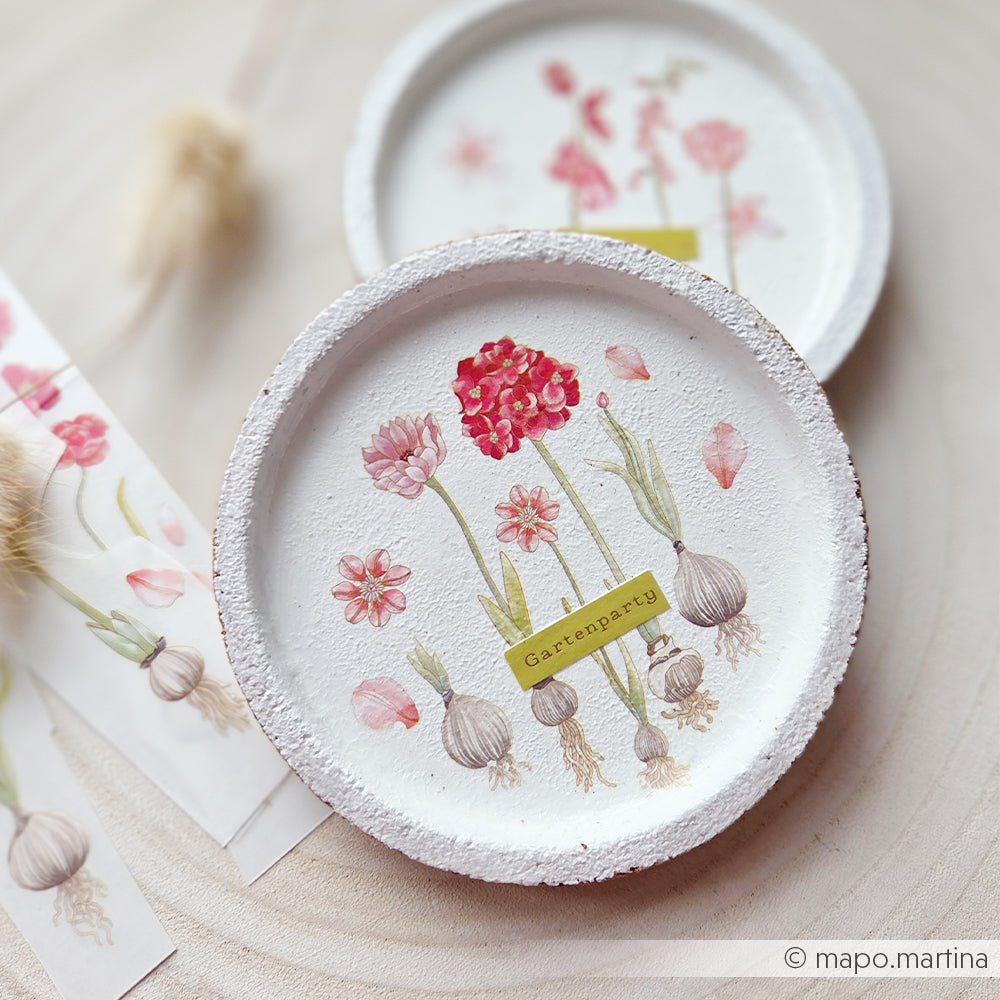 Runder weißer Teller von Alexandra Renke mit aufgemalten Blumen, Blumenzwiebeln und einem gelben Etikett mit der Aufschrift „Gartenparty“ in der Mitte – perfekt für Bastelprojekte oder um Ihrem nächsten kreativen Projekt einen Hauch von „Rub-On Gartenzauber“ zu verleihen.