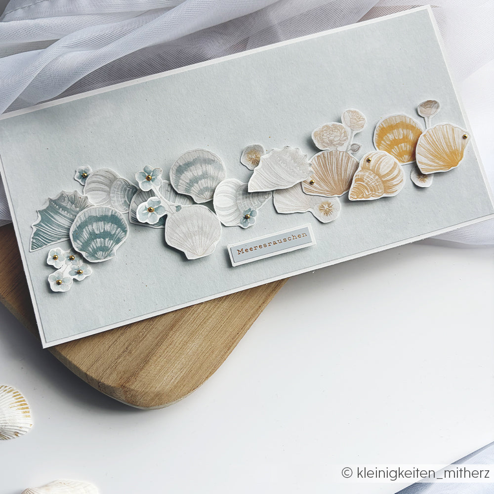 Handgefertigte Karte, verziert mit pastellfarbenen Muscheln und kleinen Blumen, mit Alexandra Renke Rub-On Meeresbrise-Akzenten, ausgestellt auf einem Holzbrett.