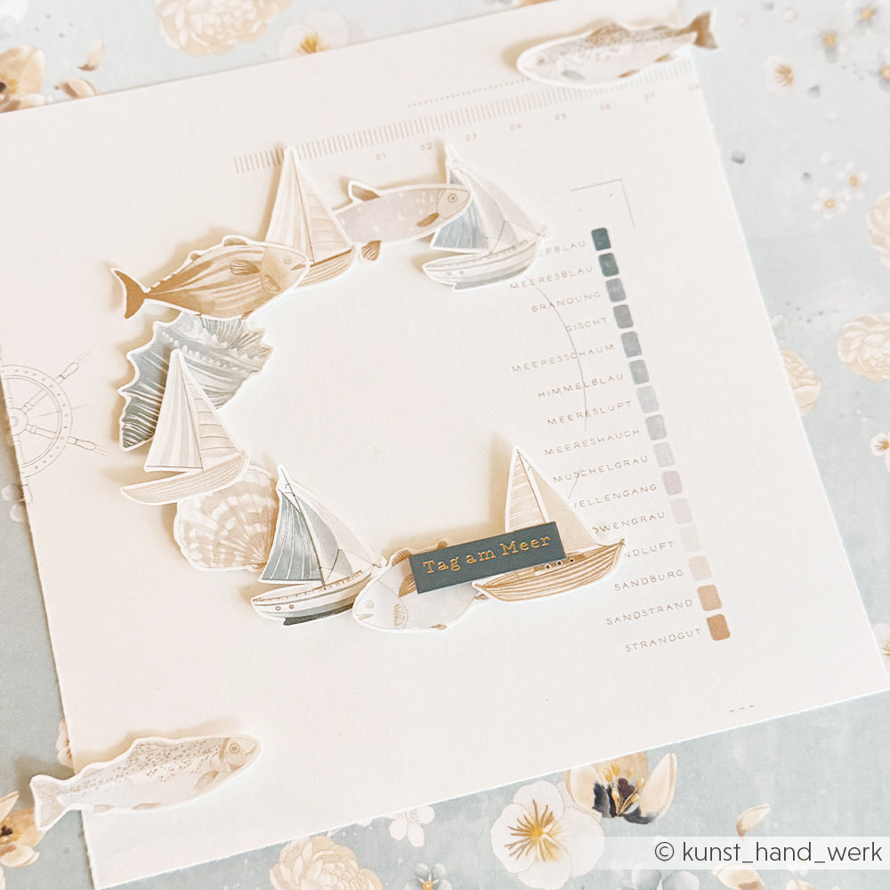 Quadratische Karte von Alexandra Renke, verziert mit Papierbooten und Fischen und mit der Aufschrift "Tag am Meer". Mit Rub-On Meeresbrise-Akzenten - perfekt für kreative, vom Meer inspirierte Bastelprojekte.