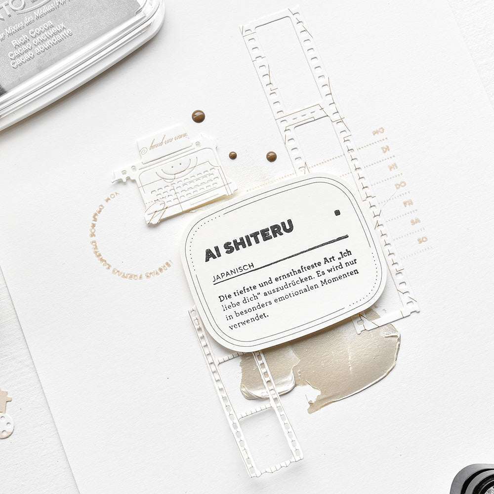 Nahaufnahme einer Karte von Alexandra Renke mit der Aufschrift "AI SHITERU" und deutschem Text, verziert mit Filmstreifen, neutralen Akzenten und einem klaren Stempeldetail für zusätzlichen Charme.