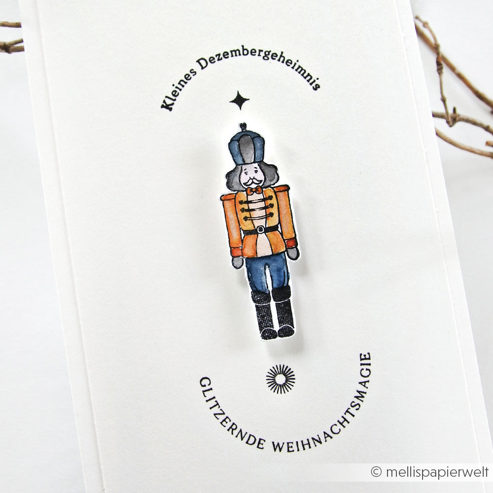 Eine handgezeichnete Nussknacker-Anstecknadel auf einer weißen Karte mit deutschem Feiertagstext, dekorativen Akzenten und Alexandra Renke Photopolymer-Stempeldetails.