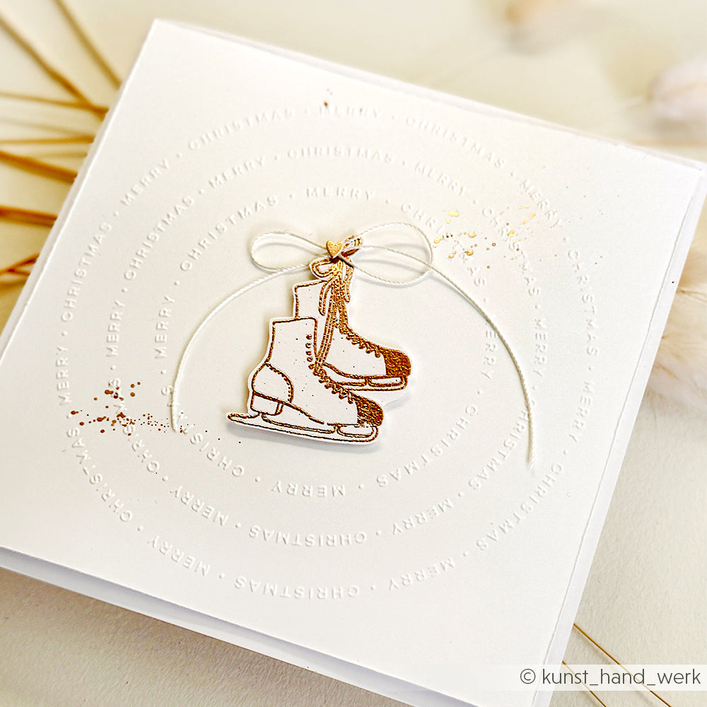 Weiße Karte von Alexandra Renke mit geprägtem "Frohe Weihnachten", goldenen Schlittschuhanhängern, die in der Mitte mit einer Schleife gebunden sind, und einem dezenten Kuschelzeit-Klarsichtstempel für einen besonders festlichen Touch.