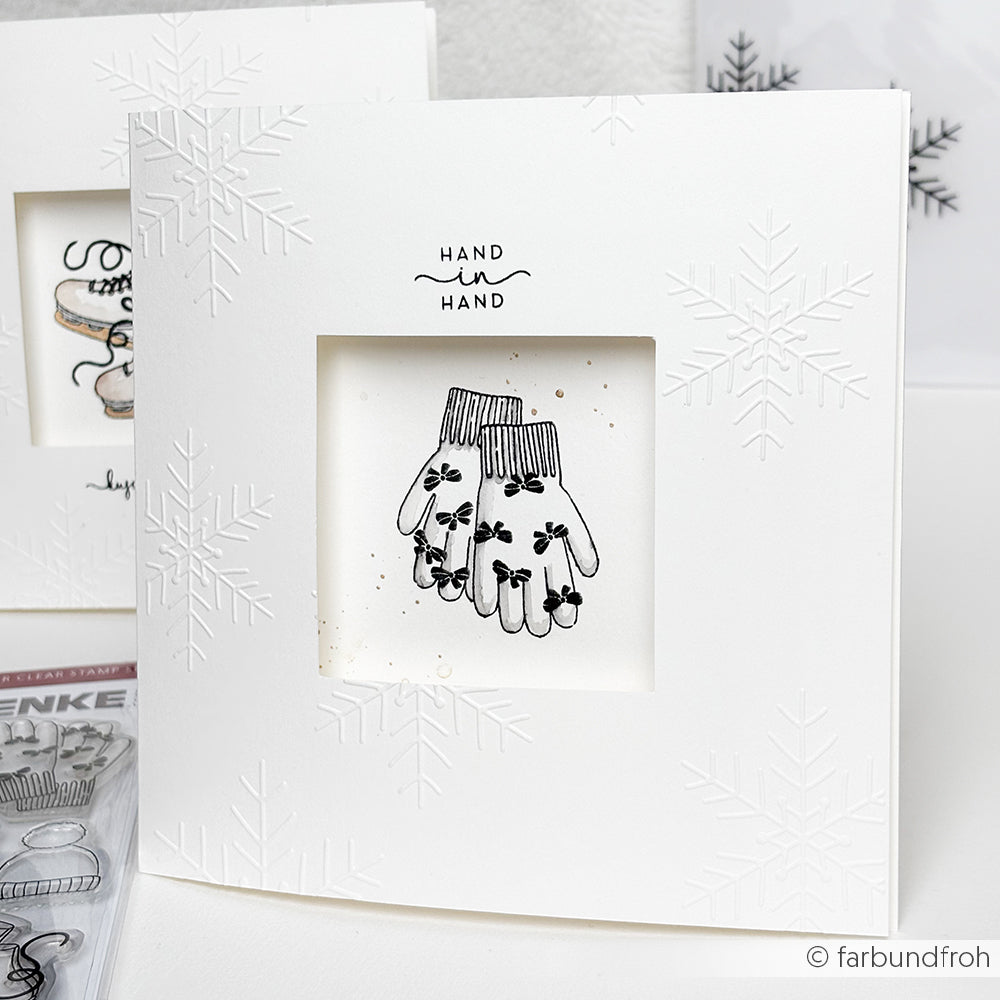 Weiße Grußkarte von Alexandra Renke mit geprägten Schneeflocken, skizzierten Fäustlingen und "Hand in Hand" darüber. Mit Photopolymer-Stempel-Details für elegante Akzente.