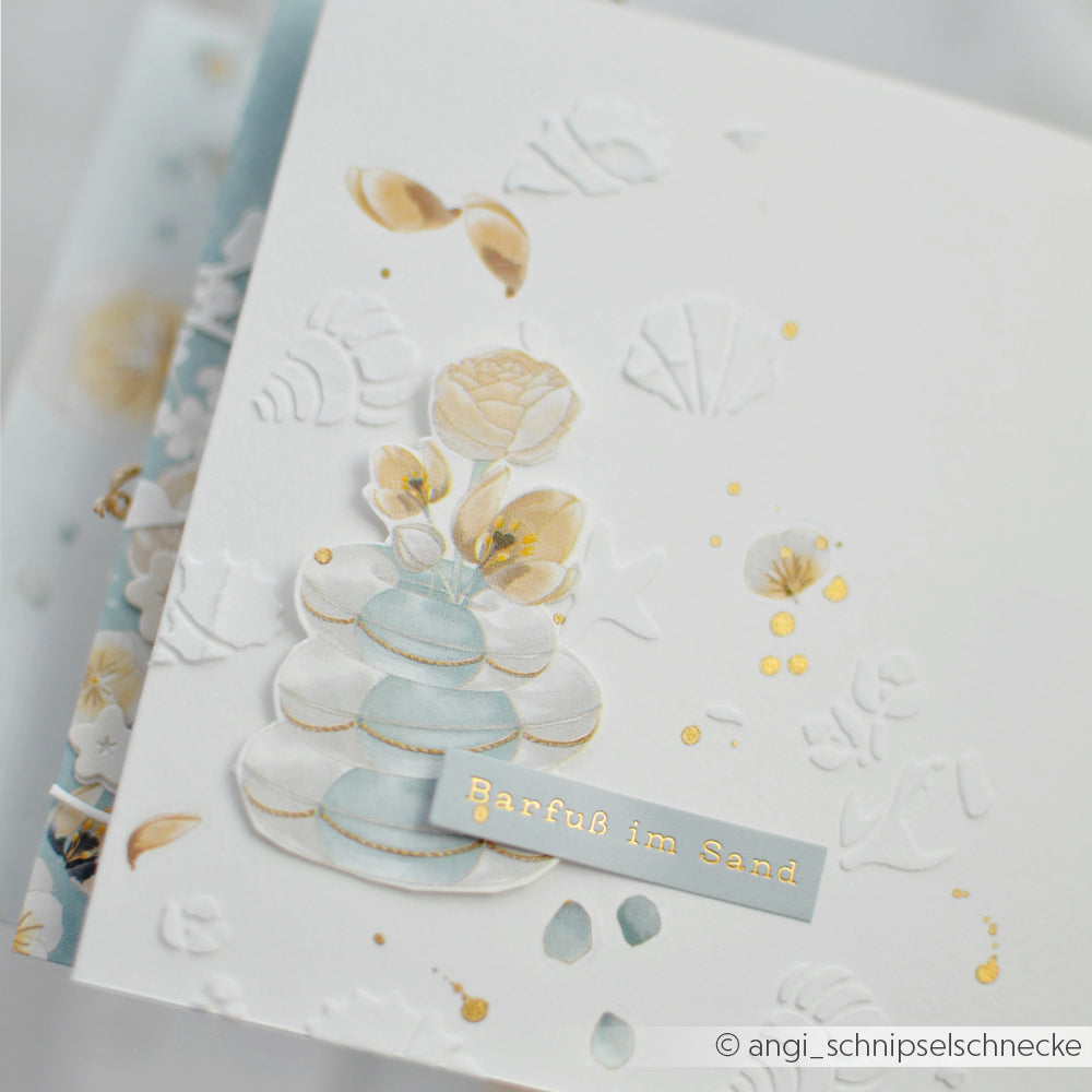 Weiße Karte mit geprägten Muscheln, goldenen Details und aufeinandergelegten Steinen, gestaltet mit der Schablone „Florale Muscheln“ von Alexandra Renke. Text: Barfuß im Sand.