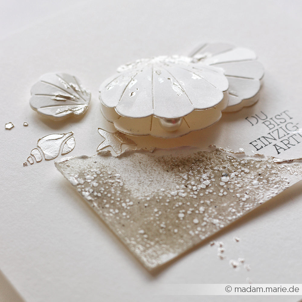 Handgefertigte Karte in Weiß und Silber mit einer mehrschichtigen Blume, Muscheln und einem glitzernden, strukturierten Quadrat auf weißem Hintergrund – ideal für Schablonen-Papierprojekte mit der Schablone „Florale Muscheln“ oder andere kreative Anwendungen von wiederverwendbaren Schablonen.