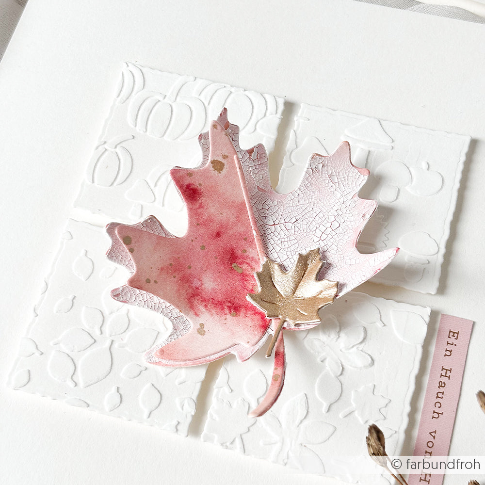 Eine strukturierte Karte mit rotem Ahornblatt, geprägten weißen Quadraten und einem kleinen goldenen Blatt darauf, gestaltet mit den „wiederverwendbar“-Schablonen „Herbstfreuden“ für zusätzliche künstlerische Akzente.