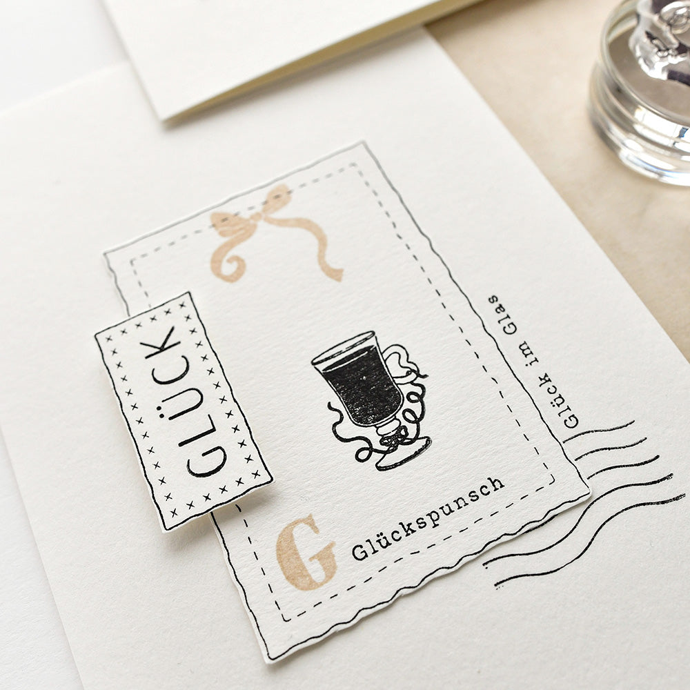 Eine Karte von Alexandra Renke zeigt eine schwarze Illustration eines Glasbechers, das deutsche Wort "Glück" auf einem kleinen Etikett und eine Photopolymer-Stempelverzierung.