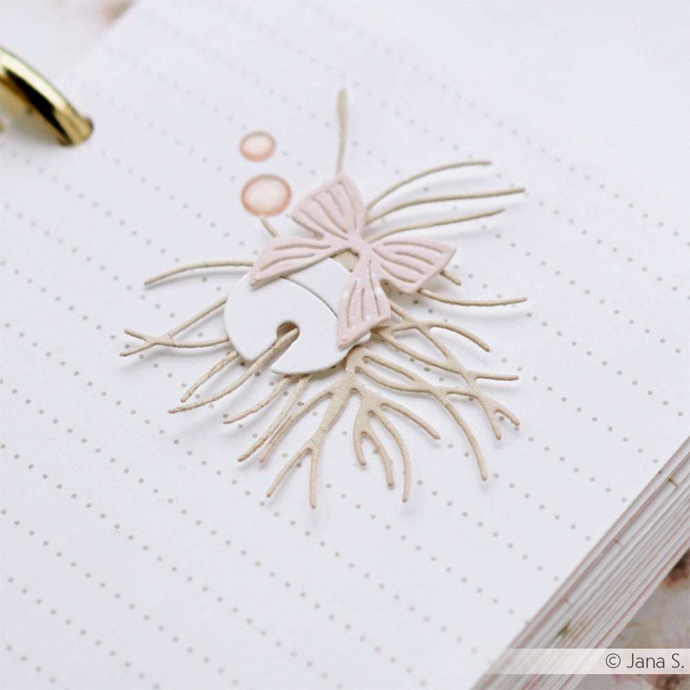 Eine gestanzte Papierverzierung mit einer Schleife und Zweigen von Alexandra Renke, arrangiert auf gepunktetem Designpapier, setzt einen schönen Akzent im Planer.