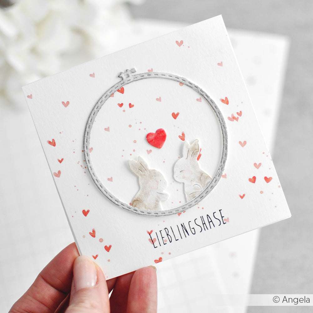 Die Hand hält eine Karte mit zwei Kaninchen, einem roten Herz und kleinen Herzen mit der Aufschrift "Lieblingshase". Hergestellt mit Alexandra Renke Designpapieren und veredelt mit der Motivstanze Stickrahmen Set Groß für einen charmanten Look.