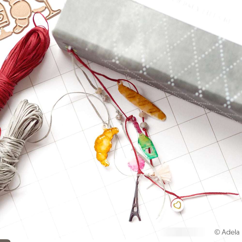 Verschiedene Anhänger und Fäden liegen neben einer gemusterten grauen Schachtel auf einem weißen Raster. Die Designpapiere und DieCut Systeme von Alexandra Renke verleihen dem Arrangement kreatives Flair.