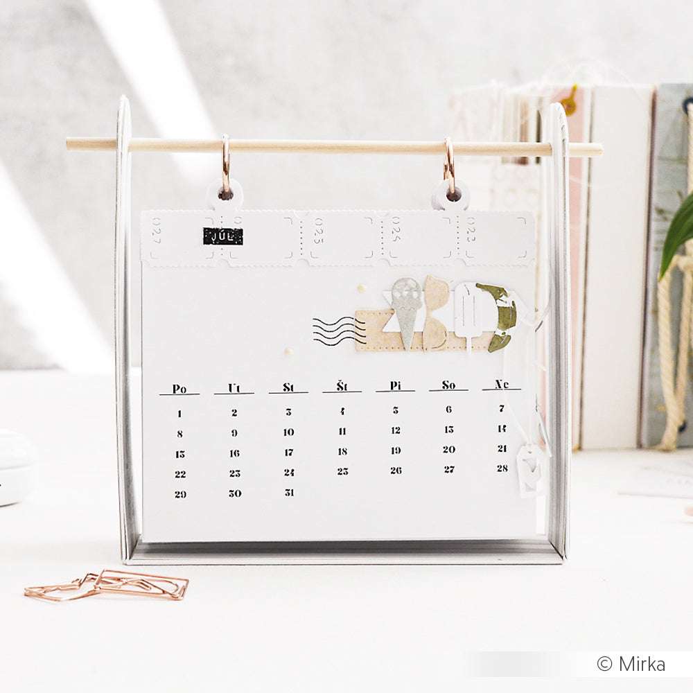 Ein minimalistischer Alexandra Renke Tischkalender für Juli hängt auf einem Ständer, umgeben von Büroklammern, Büchern und einer SWEETS Icon Set Motivstanze.
