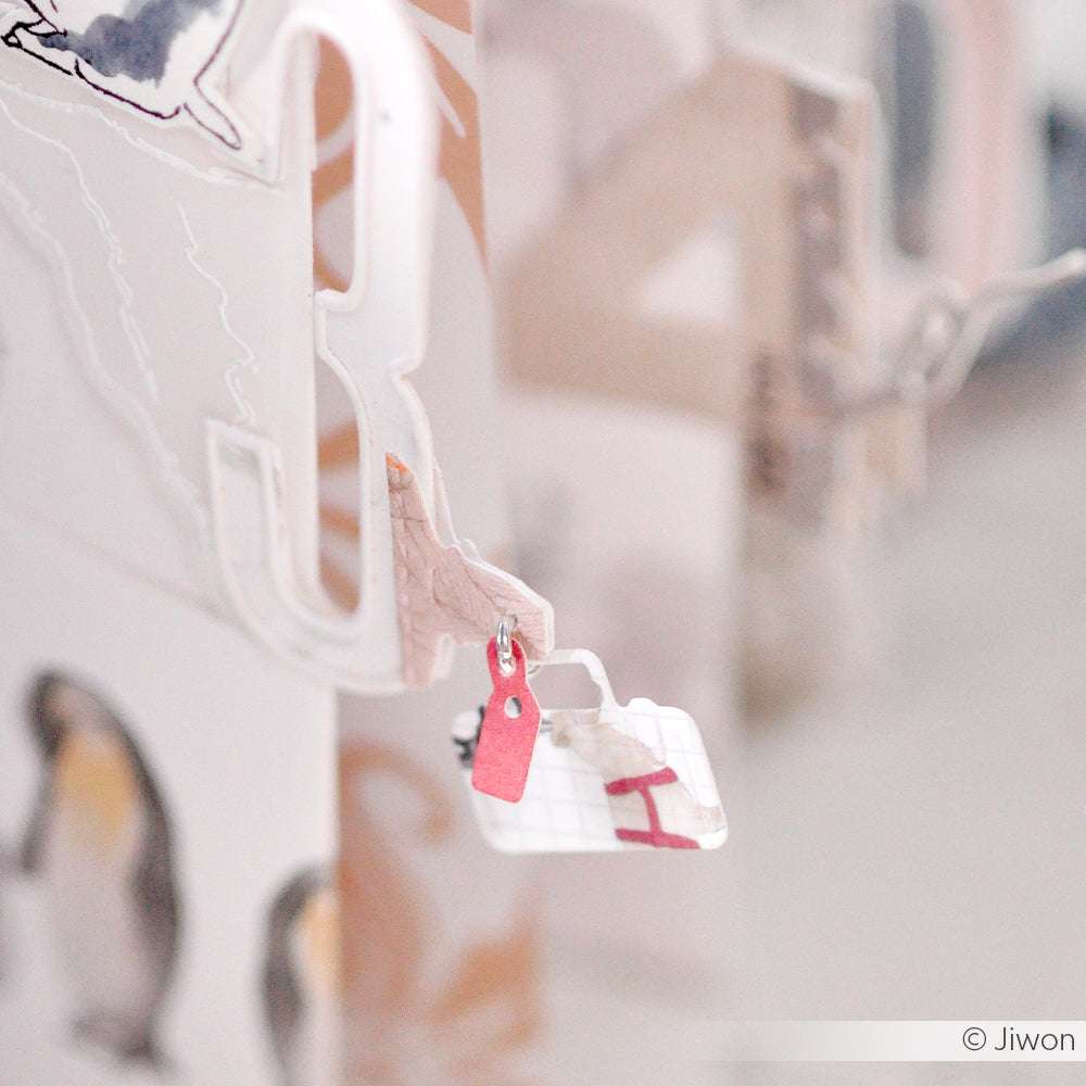 Nahaufnahme von geschichteten Papierausschnitten, die mit den Motivstanze Drei kleine Koffer Dies von Alexandra Renke hergestellt wurden, mit einem kleinen roten Anhänger mit der Aufschrift "I". Ideal für Fans von Stanzsystemen, die einzigartige Details suchen.