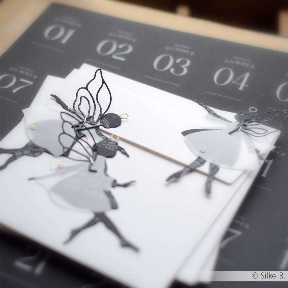 Handgemachte Feenausschnitte auf weißen Karten, hergestellt mit Alexandra Renke Motivstanze DieCut Systeme, vor einem dunklen Kalenderblatt-Hintergrund.