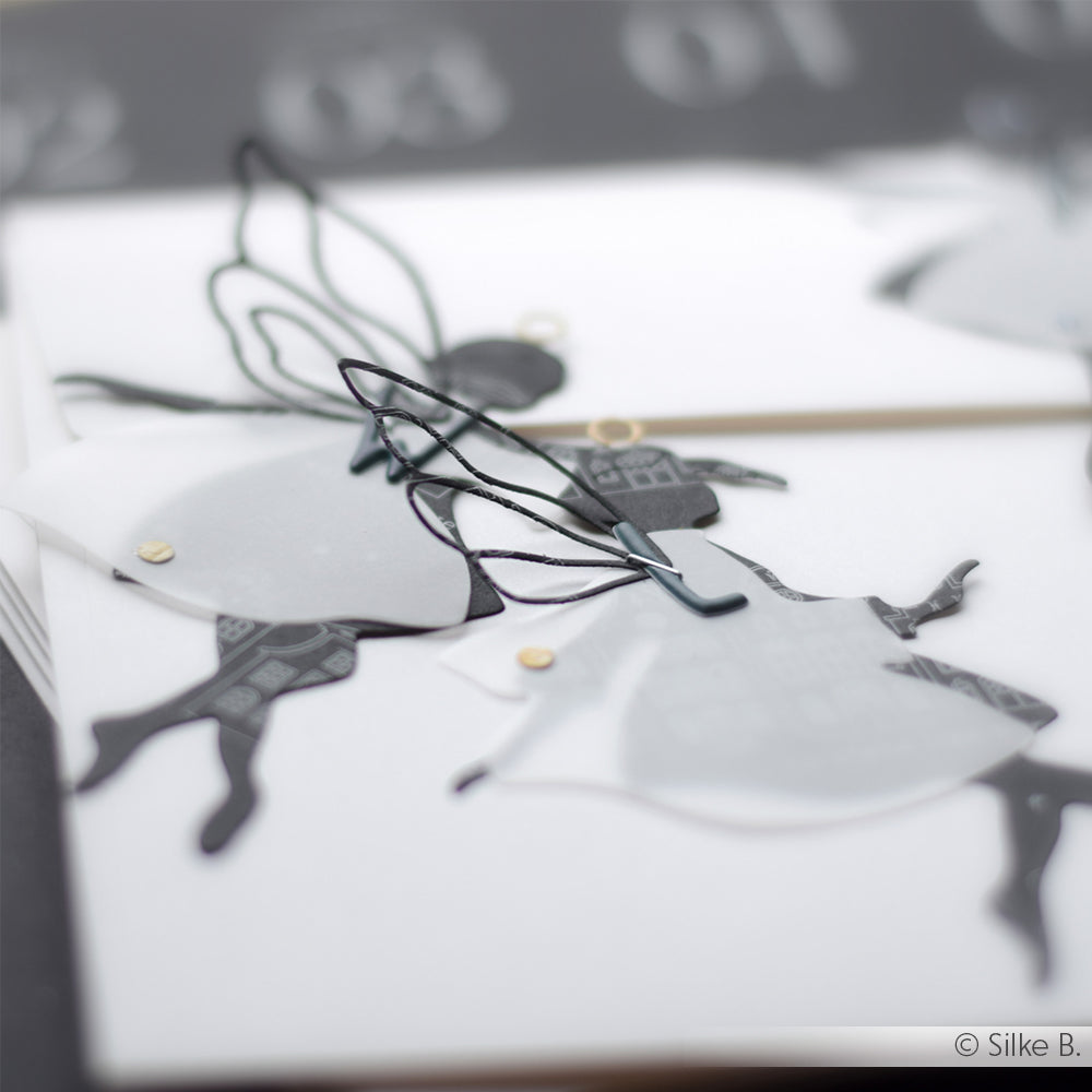 Nahaufnahme von zwei schwarz-weißen Feenornamenten aus Papier mit durchscheinenden Flügeln, hergestellt mit der Alexandra Renke Motivstanze Tanz Fee Lala, ausgestellt auf einer weißen Fläche.