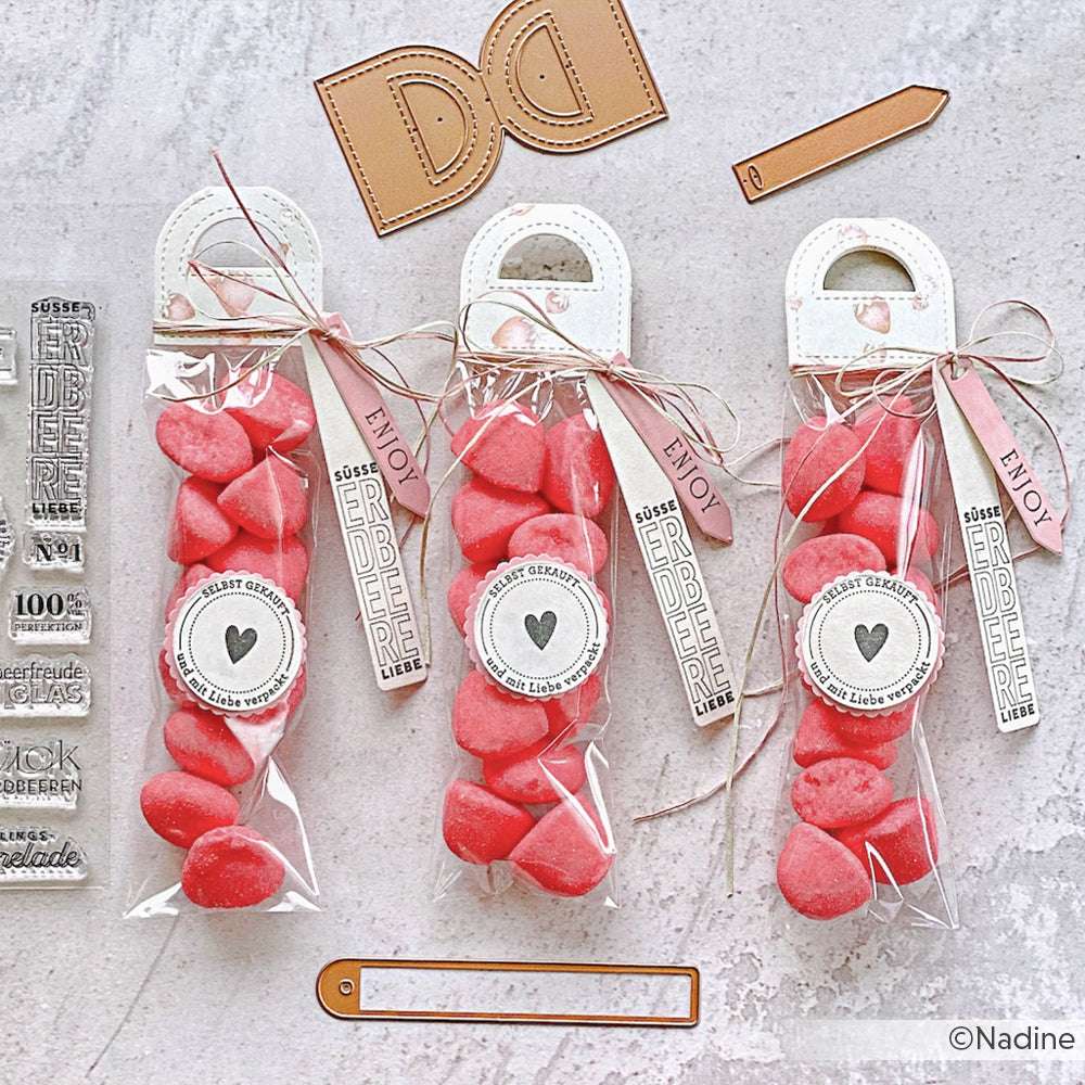 Vier durchsichtige Tüten, gefüllt mit roten, herzförmigen Erdbeertraum-Bonbons von Alexandra Renke, verschnürt mit Schnur und Anhängern, stehen auf einem hellen, strukturierten Hintergrund.