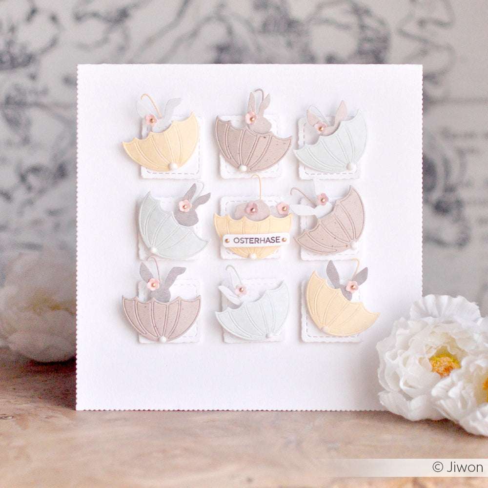 Eine weiße Karte von Alexandra Renke zeigt pastellfarbene Regenschirm- und Hasenmotive in einem Raster, mit dem Osterhasen in der Mitte, gestaltet mit Designpapieren und der Motivstanze Regenschirm.